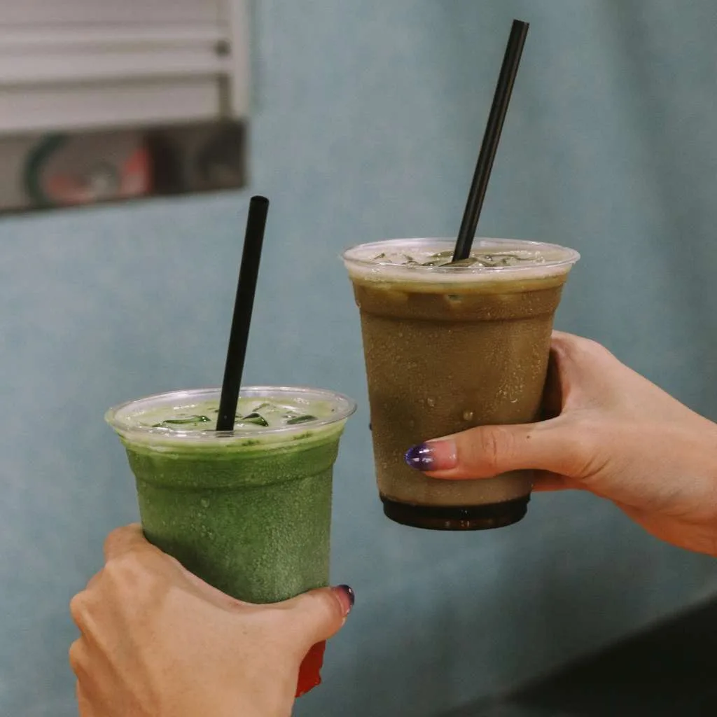 An iced matcha and an iced dirty matcha from cornershop held up side by side against the pale blue wall outside the stall