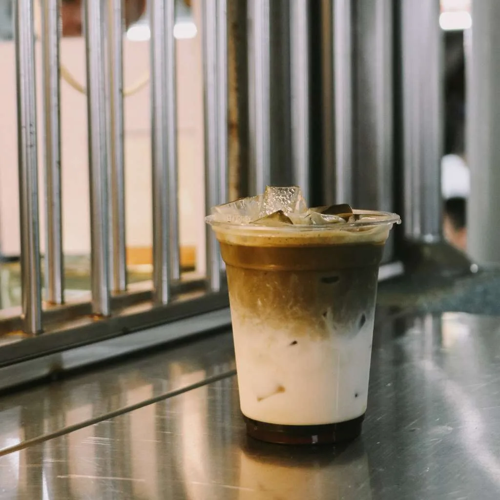 cornershop's iced maple houjicha in a clear cup on the stall's stainless ledge, roasted-tea brown settling over a milk base, hawker-centre railings behind