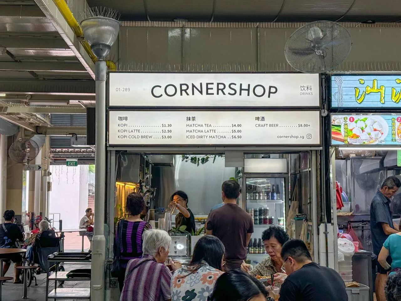 The lit CORNERSHOP lightbox sign and stall front seen from across the walkway at Marine Terrace Market, with hawker tables in the foreground