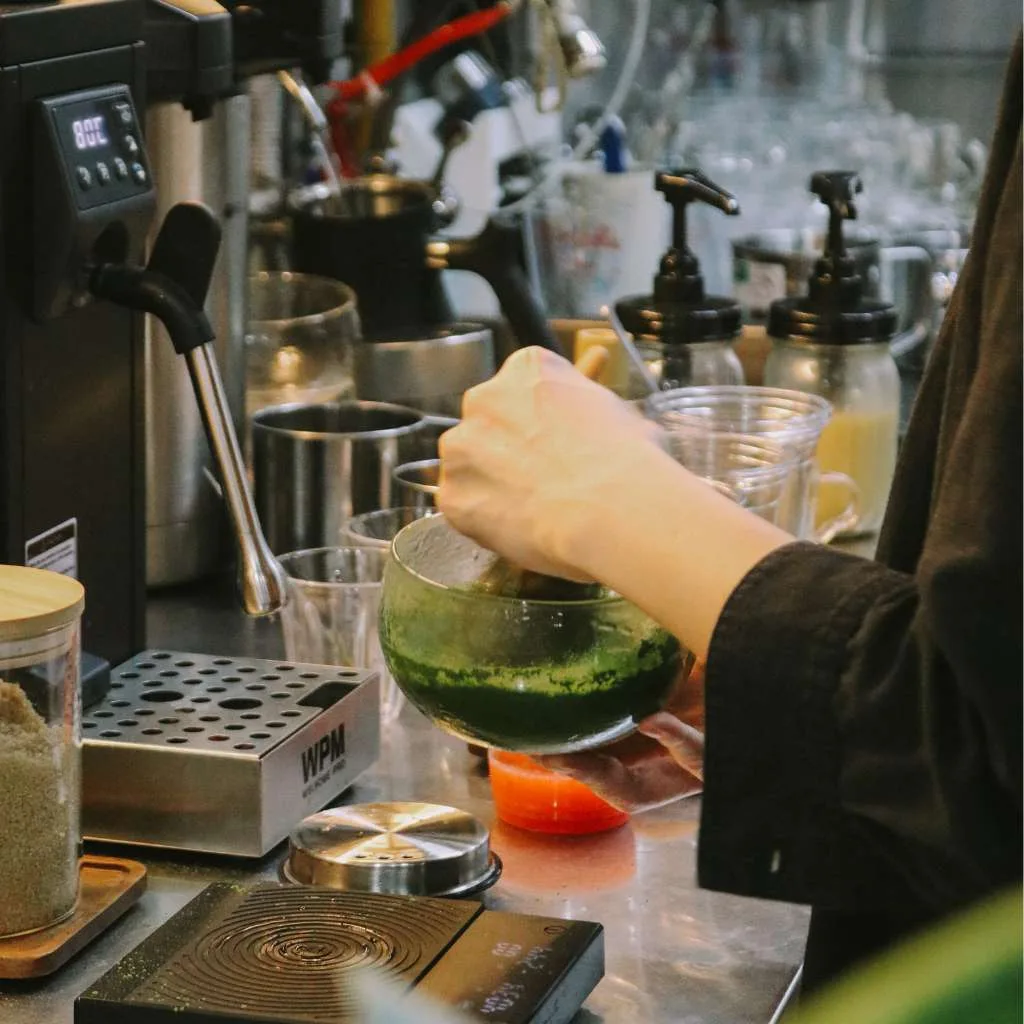 A hand whisking bright green matcha in a glass chawan on cornershop's stainless counter, beside a temperature-controlled hot-water tower and syrup pumps